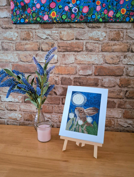 Artwork of a rabbit with flowers on an easel against a brick wall with a colorful floral painting above.